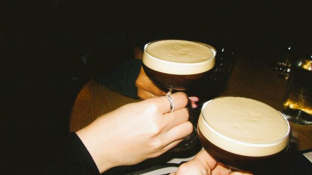 Two people clinking glasses filled with a creamy, frothy drink in a dimly lit setting.