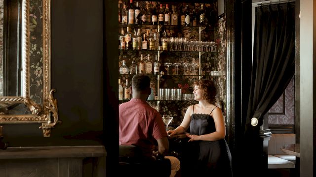 A couple is seated at a dimly lit bar, surrounded by shelves of bottles, in an elegant room with a fireplace and a large mirror on the wall.