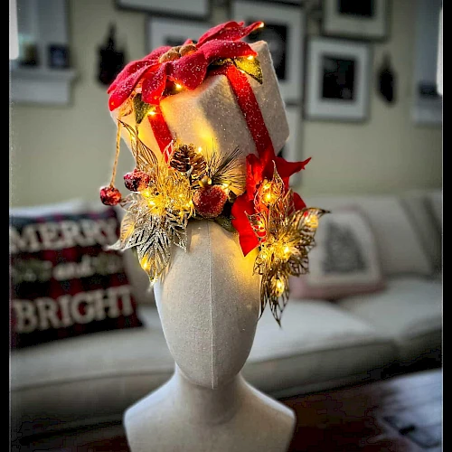 A wrapped gift hat adorned with red ribbons, gold ornaments, and warm lights on a mannequin head, festive decor in the background.