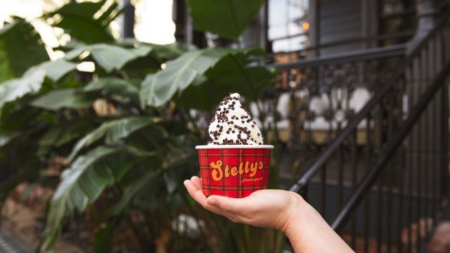A hand holds a red cup labeled "Bielly" topped with a generous swirl of cookies and cream ice cream, outdoors with green plants in the background.