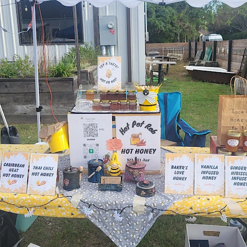 A table outdoors with many labeled boxes and bottles of hot honey gifts, jars, and a small display board under a canopy.