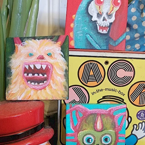 Colorful, hand-painted monster portraits on blocks, featuring fanged faces, big eyes, and quirky details; stacked with patterned boxes and jars nearby.