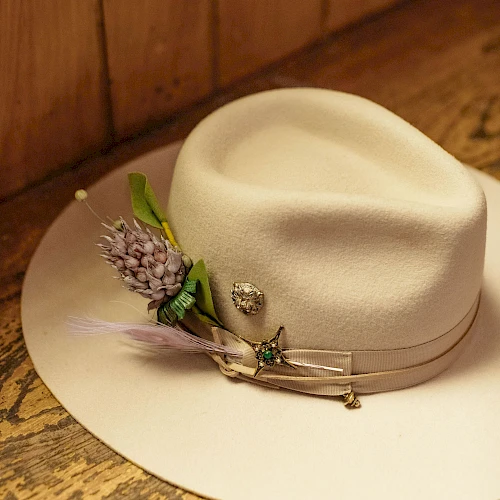 A cream fedora with a pale band sits on a wooden surface, adorned with a purple flower boutonniere and a few delicate pins.