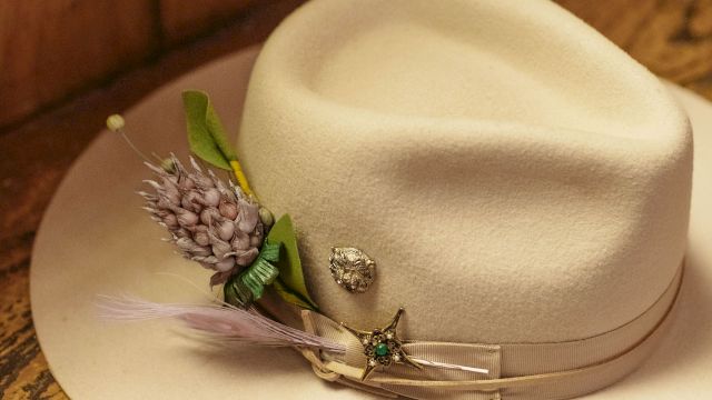 A cream fedora with a pale band sits on a wooden surface, adorned with a purple flower boutonniere and a few delicate pins.