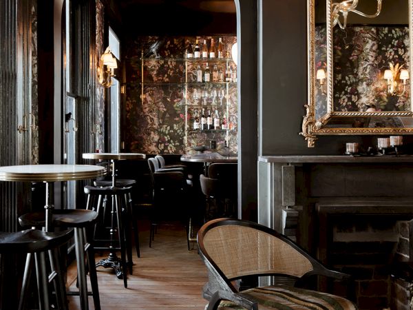 A moody, vintage-style bar or lounge with dark walls, arched doorway, tall stools at a high counter, ornate mirror, and a painted wood chair in a patterned rug.