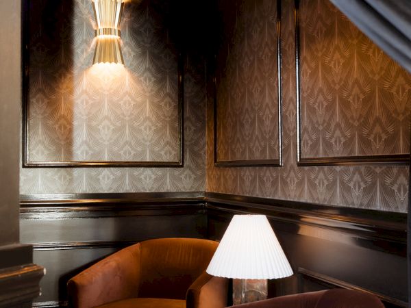 A cozy, dimly lit lounge with two brown chairs, a small table lamp, patterned wallpaper, and a hanging ceiling light. End.