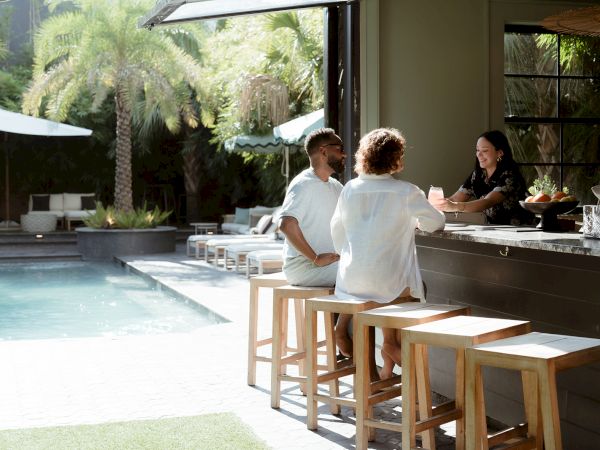 A poolside bar scene with three people at a counter, outdoor lounge area, and tropical greenery in the background, sharing drinks.