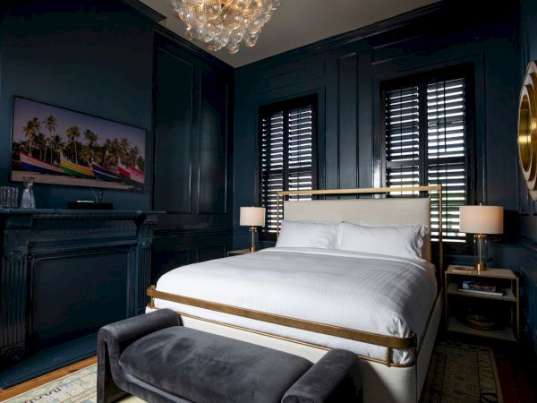 A luxurious bedroom with dark blue paneled walls, a white bed, matching nightstands, two lamps, a bench, chandelier, and a wall-mounted TV.