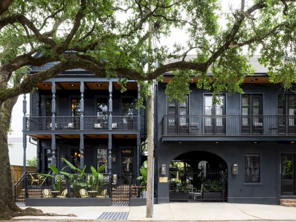 A two-story dark blue building with a wraparound porch, balconies, and arches, shaded by large trees along a sidewalk.