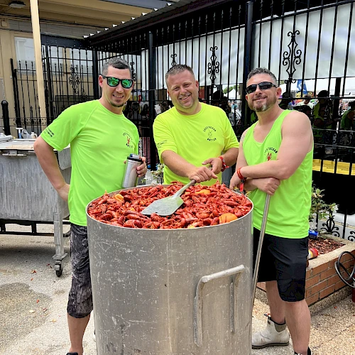 Three men in neon shirts stir a giant pot of crawfish outdoors, smiling as they cook a seafood boil.