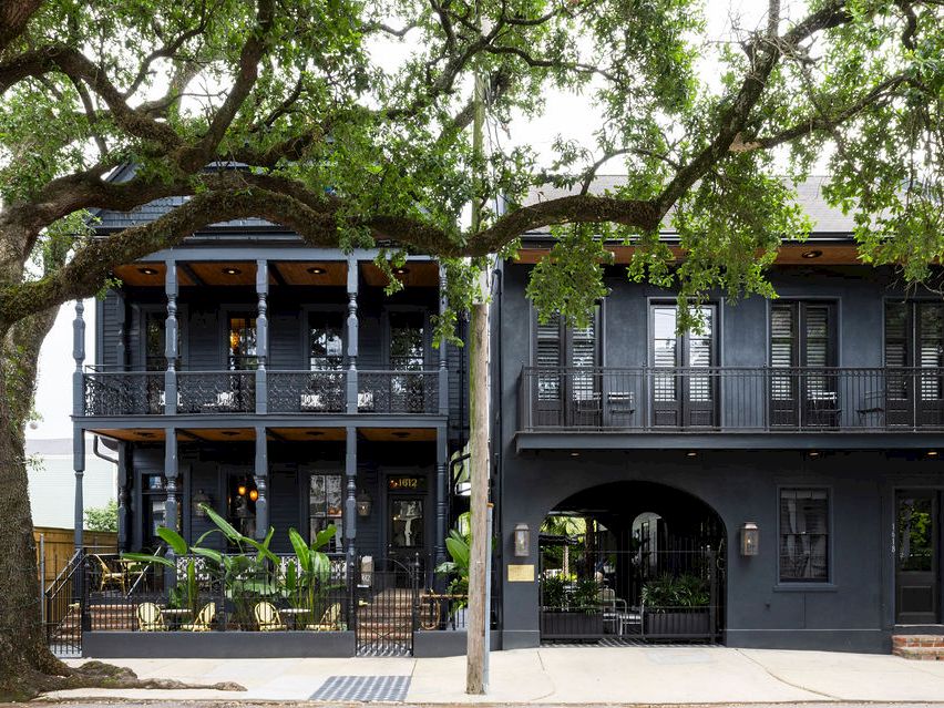 A historic-looking building with two balconies and an arched entry, framed by a large leafy tree in front.