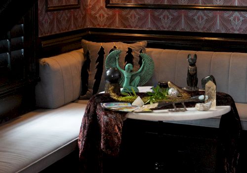 Cozy lounge with tufted couch, ornate damask wallpaper, and a side table covered with decorative statues and knickknacks.
