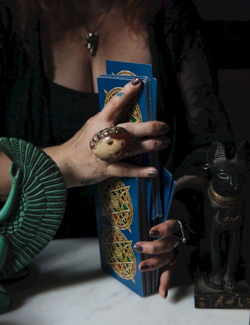 A tarot reader holds a deck of blue cards while a crystal ball and dark figurines sit on the table.