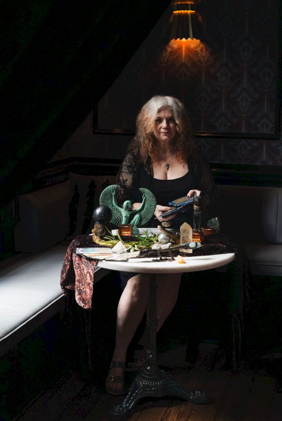 A woman sits at a candlelit table with tarot/cards, crystals, and mystical decor, facing the camera in a dark lounge.