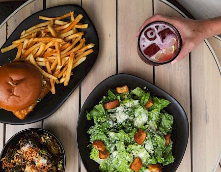 Burger and fries, Caesar salad, berry soda in a glass, and a bowl of roasted vegetables on the table.