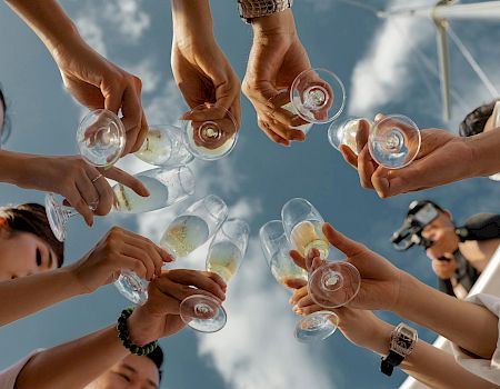 A group cheers with tiny glasses in a sunny outdoor setting, clinking drinks together as they toast one another.