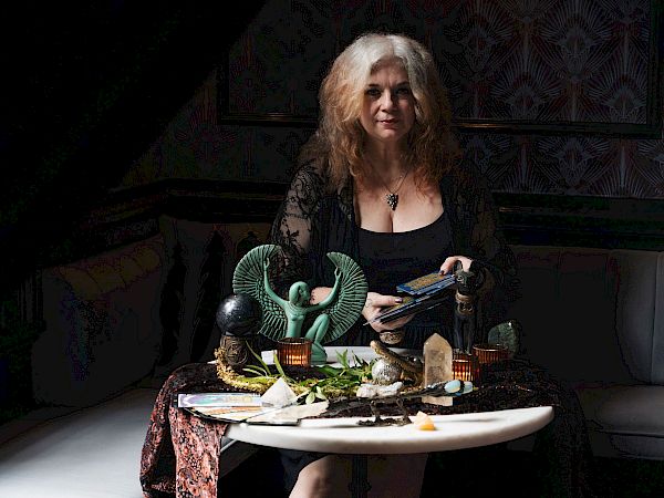 A woman sits at a table of mystical d&eacute;cor, holding tarot-style cards in a dimly lit room with patterned wallpaper.