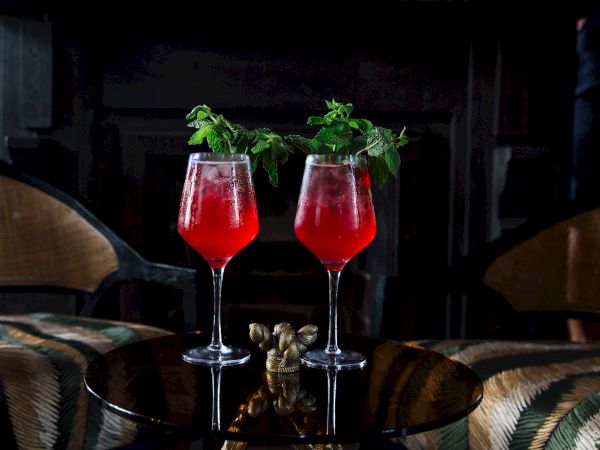 Two stemmed glasses filled with a red cocktail, topped with fresh green mint leaves, sit on a wooden table.