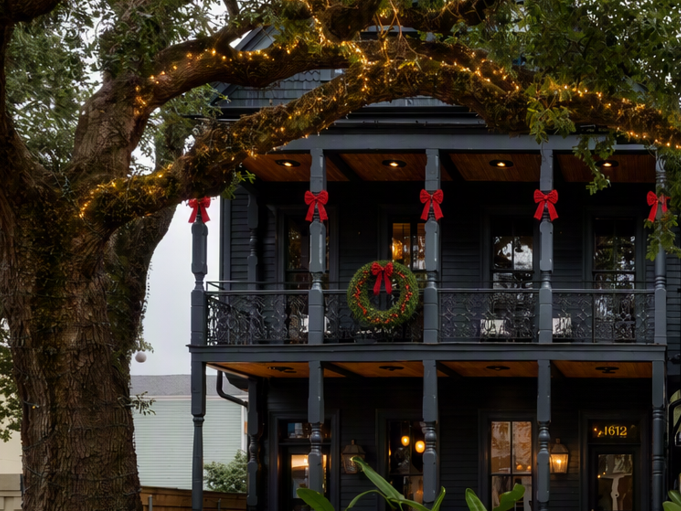 A two-story house is lit with warm string lights and red bows, with wreaths and ornaments on the porch under a tree.