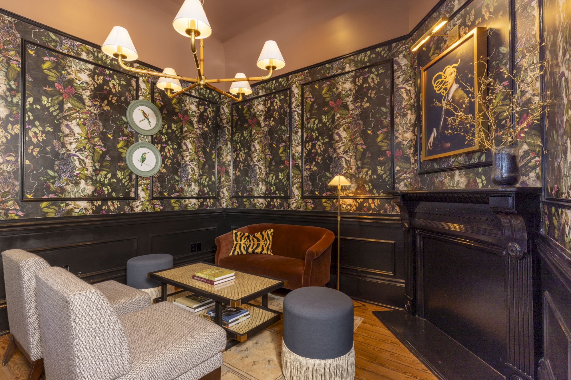 A cozy lounge with patterned floral wall panels, vintage brass light fixtures, framed art, and comfy seating in warm amber light.