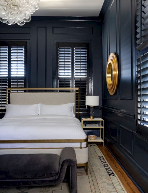 A stylish dark bedroom with a bed, wall shutters, gold accents, a chandelier, and a modern lamp in a cozy corner.