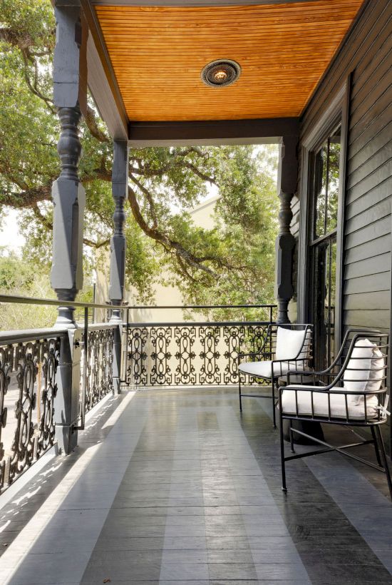 A quiet porch with a wooden ceiling, ornate metal railing, and two cushioned chairs, surrounded by leafy trees in sunlight.