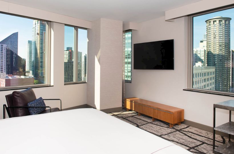 A modern room with large windows showcasing a cityscape view, a bed, TV, chair, and a side table.