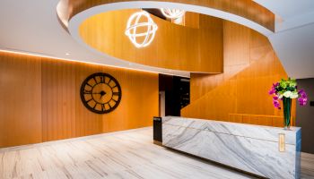 A modern lobby with a marble reception desk, wooden walls, a large clock, and ceiling lights.