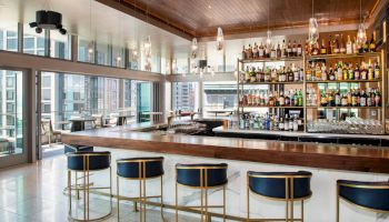 A modern bar with five stools, shelves of bottles, and large windows offering a cityscape view, creating a stylish ambiance.