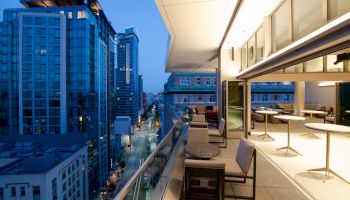 A modern cityscape view from a high-rise balcony at dusk, with tables and seating, overlooking streets and buildings under a blue sky.