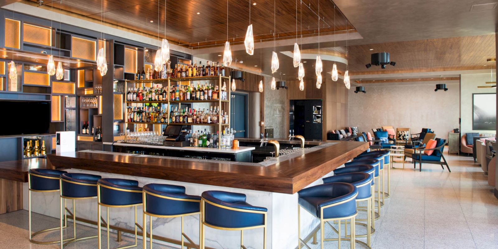 The image shows a modern bar with blue chairs, a well-stocked shelf, and hanging lights, in a spacious lounge setting.
