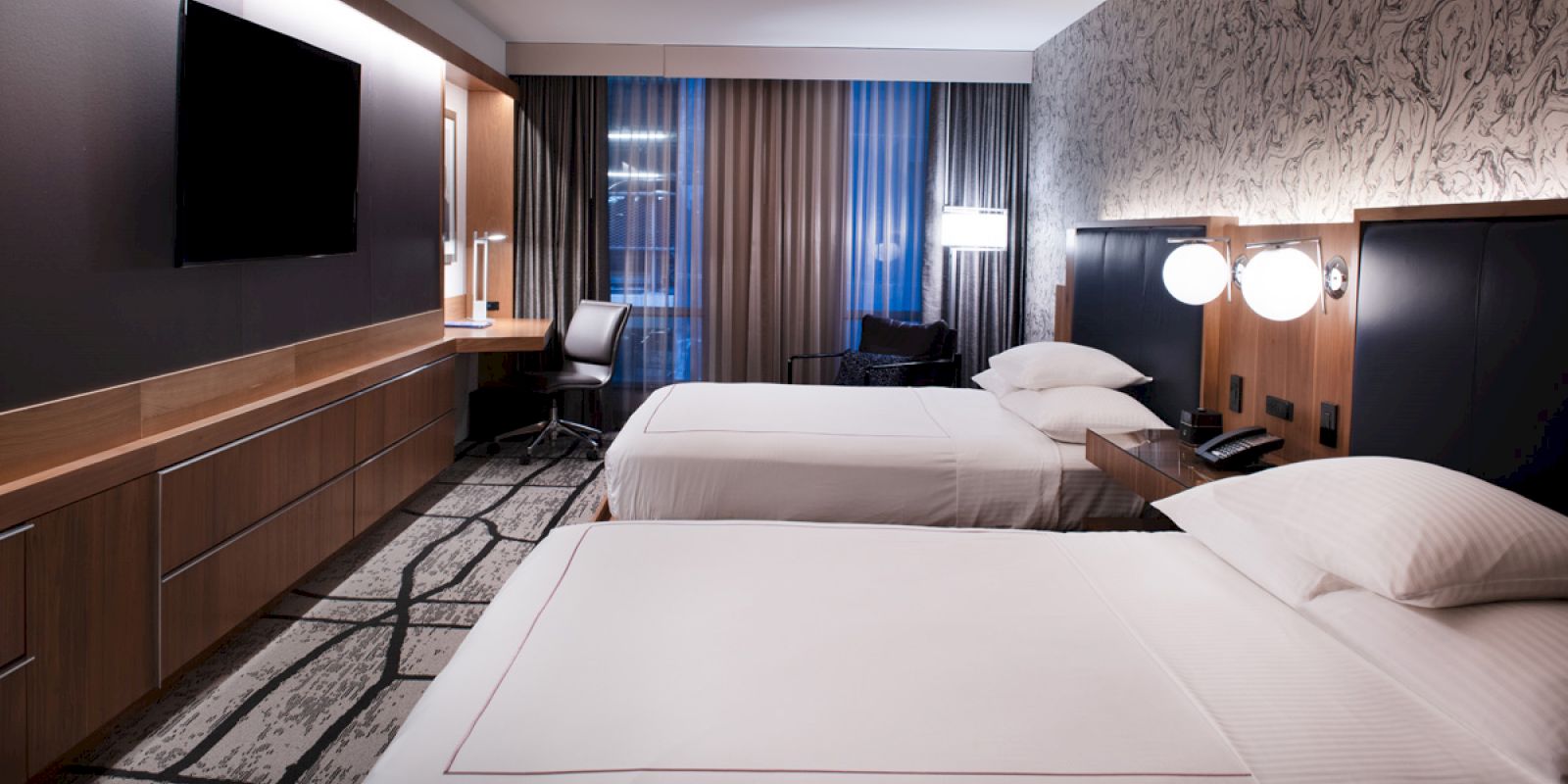 The image shows a modern hotel room with two beds, a large TV, a desk with a chair, and a stylish patterned carpet.