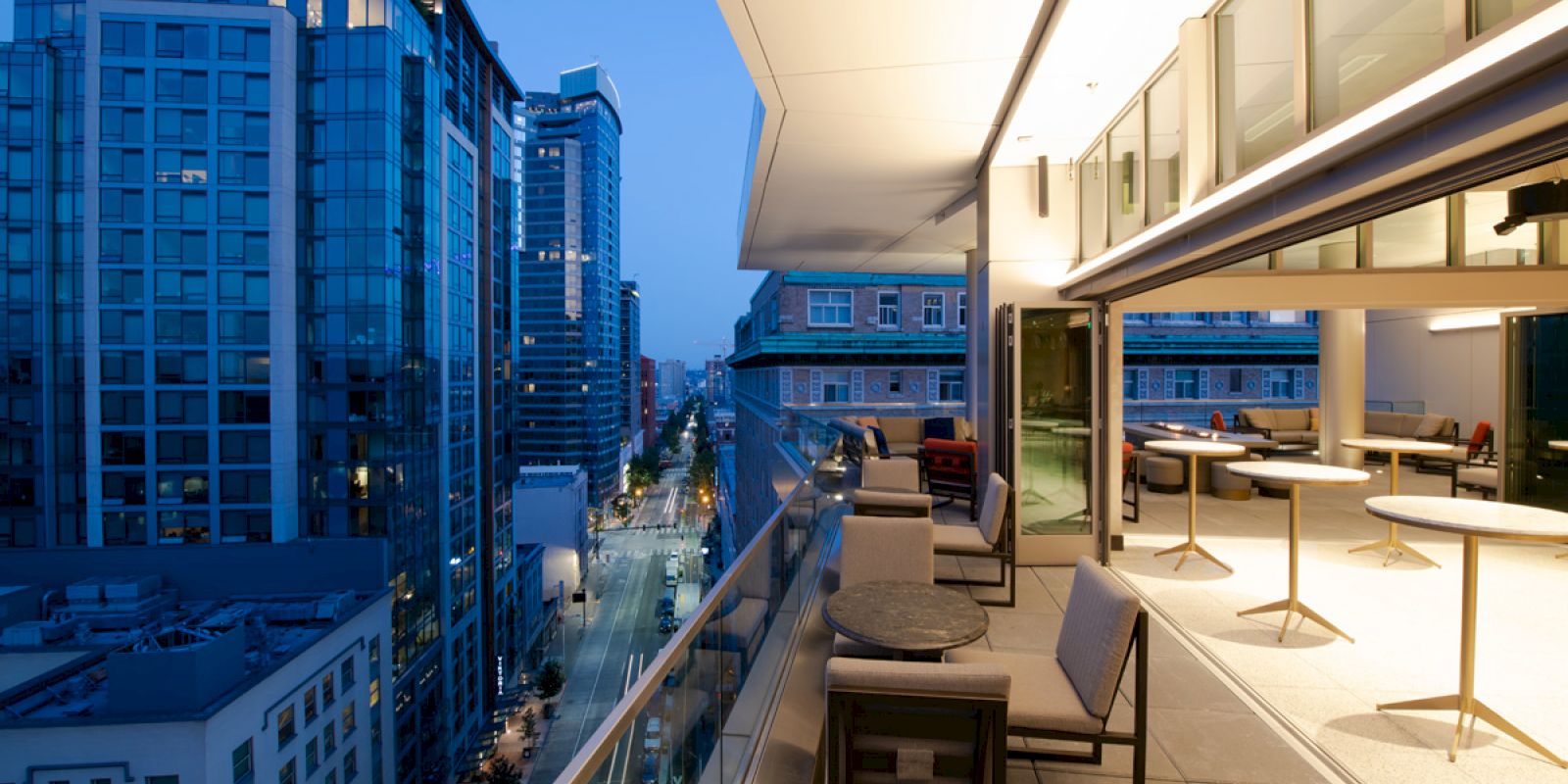 A modern rooftop terrace at twilight with cityscape views, outdoor seating, and large windows showing an interior space.