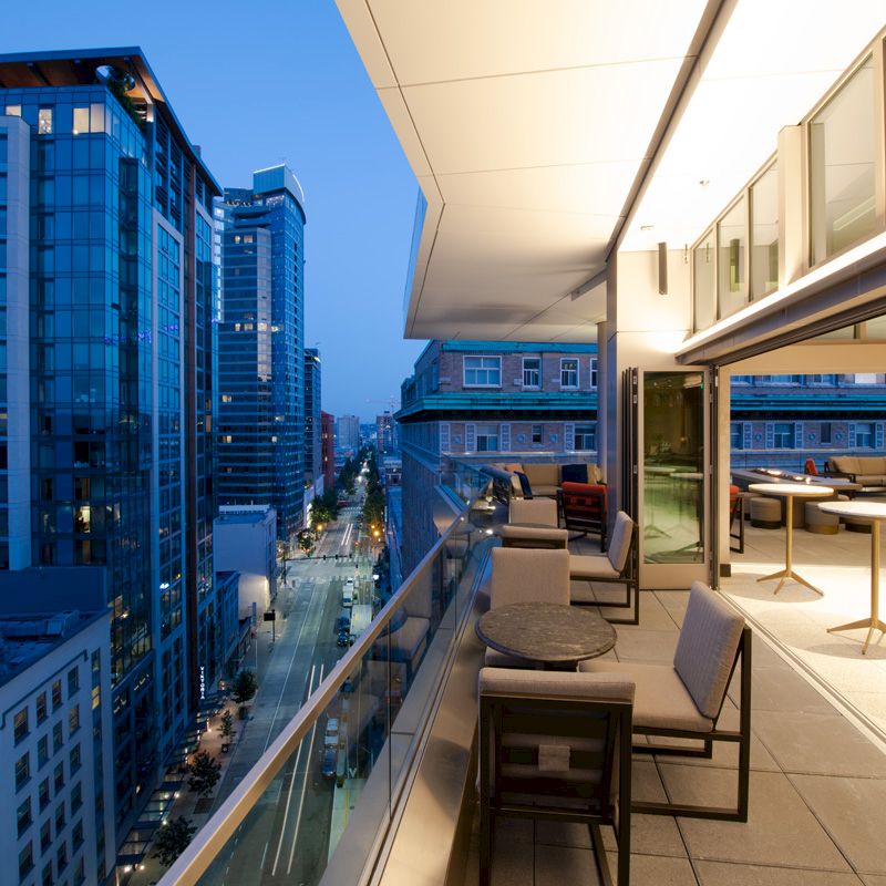 A cityscape view from a modern balcony with seating, overlooking a street lined with tall buildings at dusk.