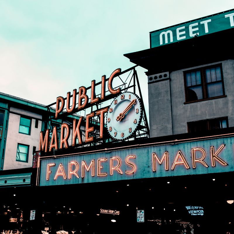 The image shows a public market sign with a clock and "Farmers Market" in neon. Buildings in the background complete the scene.