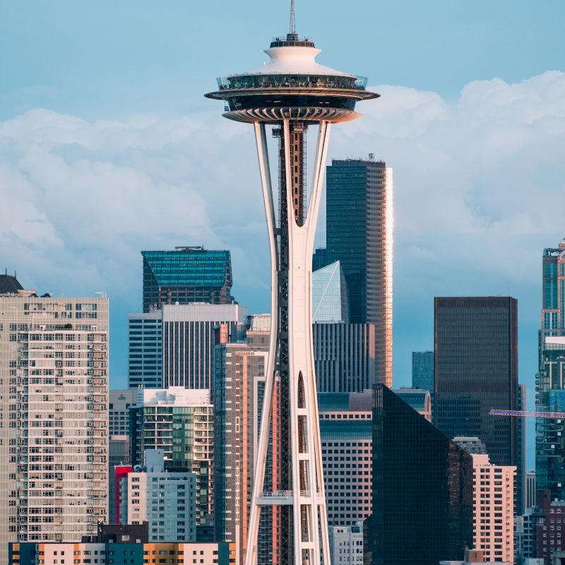 The image shows the Space Needle with a backdrop of tall buildings and a cloudy sky, characteristic of an urban cityscape.