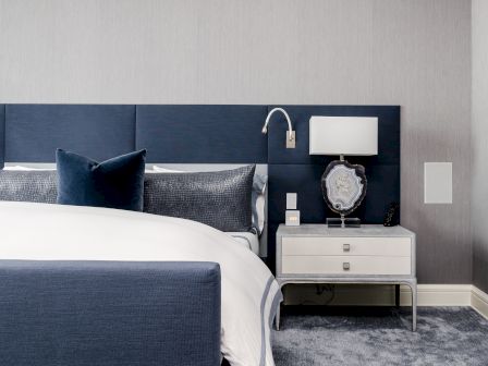 A modern bedroom with a navy bed, gray and navy pillows, a white nightstand with a lamp, and a decorative item on top.
