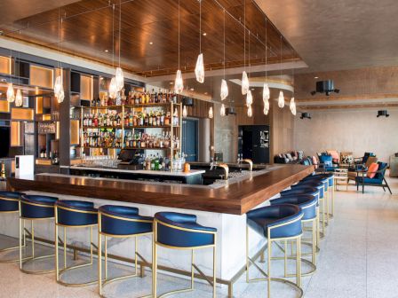 A modern bar with blue chairs, hanging lights, and a stocked liquor shelf, featuring a lounge area with seating in the background.