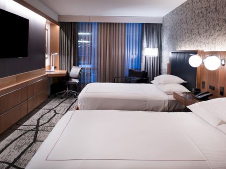 The image shows a modern hotel room with two beds, a TV, desk, and chair, featuring stylish decor and ambient lighting.