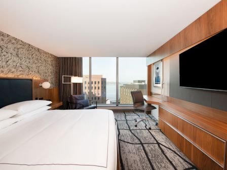 A modern hotel room with a large bed, patterned wallpaper, desk, chair, and TV, overlooking a cityscape through large windows.