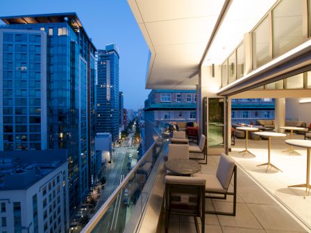 A modern city view featuring tall buildings and a stylish rooftop terrace with seating and tables, captured at dusk.