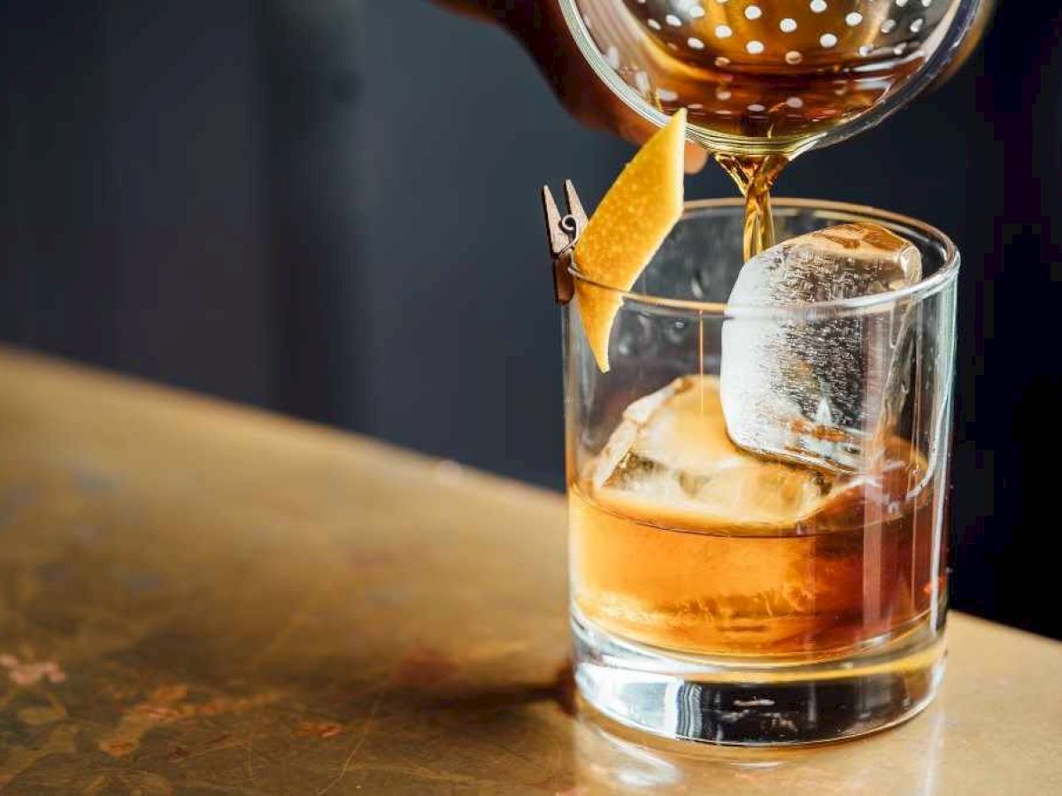 A glass with a large ice cube, being filled with a brown drink, garnished with a lemon twist on a metal bar surface.