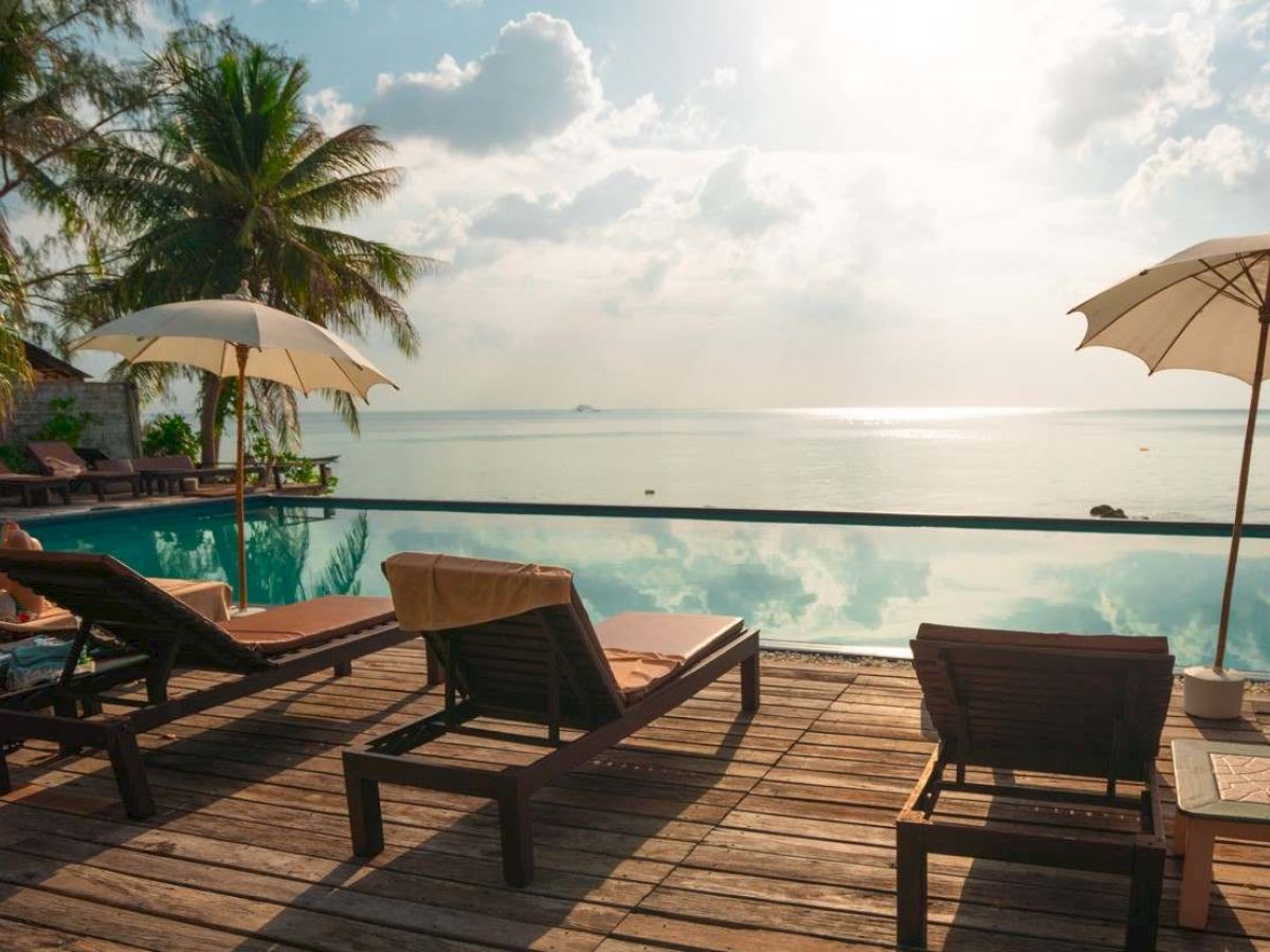 A beachfront pool scene with lounge chairs, parasols, and palm trees, overlooking a calm sea under a bright sky with scattered clouds.