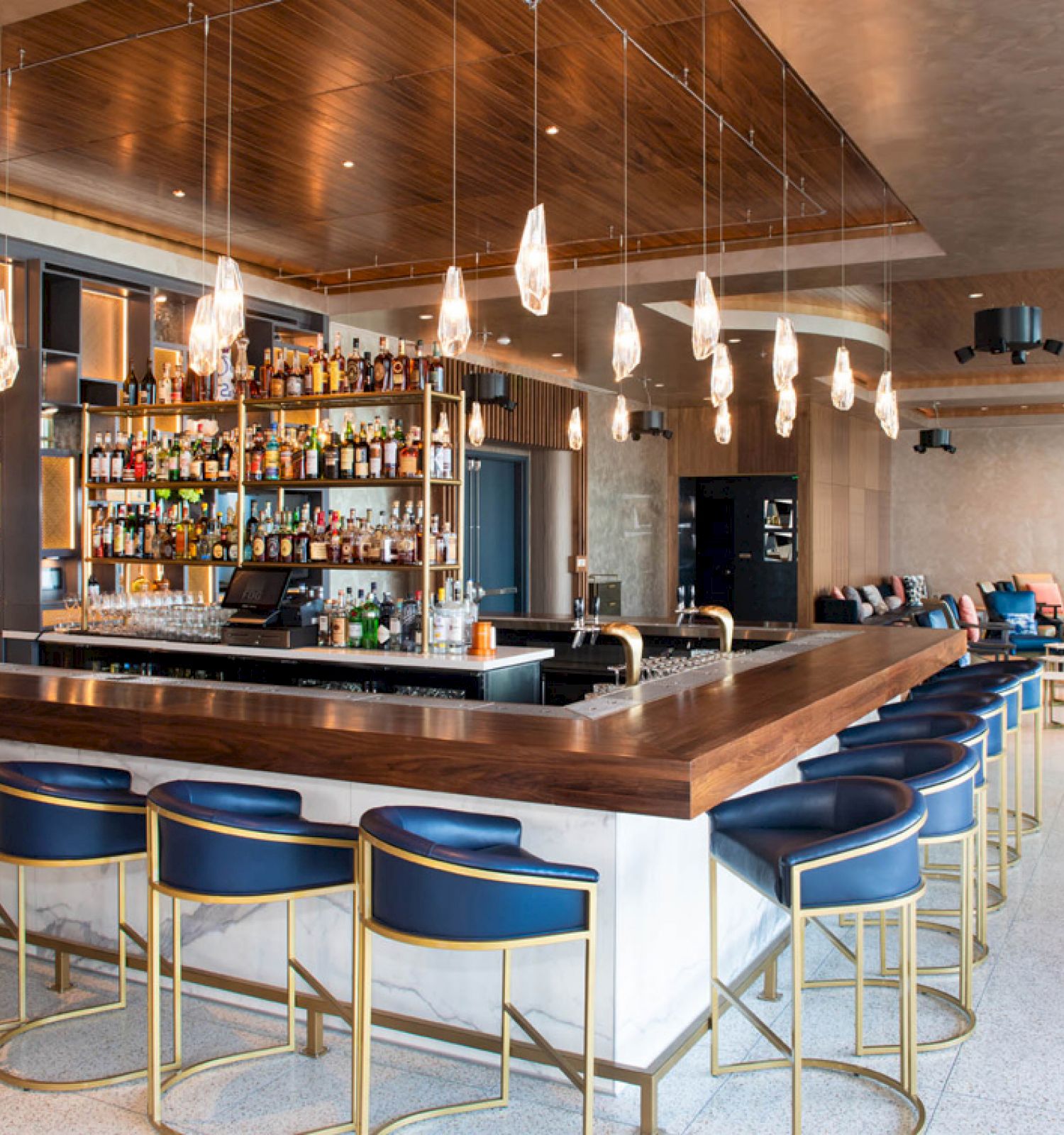 A modern bar with blue stools, overhead lights, and a fully stocked bar. There's a seating area in the background with plush chairs.