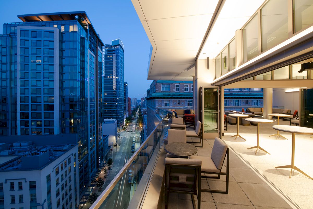 A modern cityscape at dusk with tall buildings and a rooftop terrace featuring seating and tables with a view.