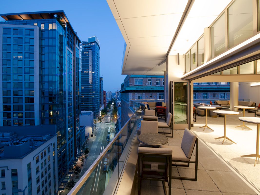 A modern cityscape at dusk with tall buildings and a rooftop terrace featuring seating and tables with a view.