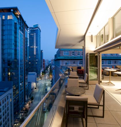 A modern city view from a balcony at twilight, with outdoor seating and illuminated interiors, overlooking tall buildings and a street.