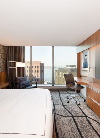 A modern hotel room with a large bed, wall-mounted TV, desk with chair, and a window view of buildings and water outside.