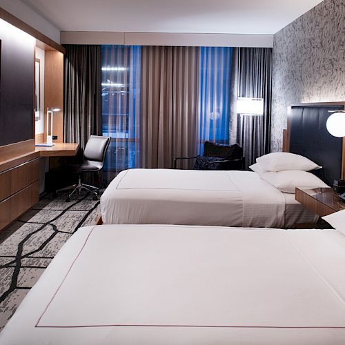 The image shows a modern hotel room with two beds, a wall-mounted TV, desk, chair, and stylish lighting, featuring a patterned wall design.