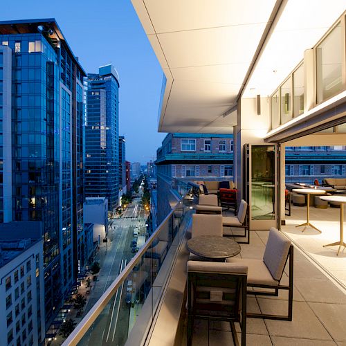 A modern rooftop patio with tables and seating overlooks a cityscape of tall buildings during twilight, featuring glass and lights.